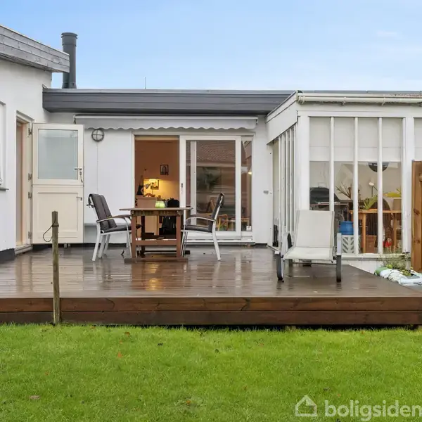 En træterrasse med et bord og to stole står udenfor et hvidt hus. Der er en dør og vinduer i baggrunden. Græsplæne omgiver terrassen.