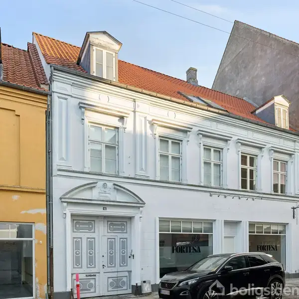 En hvid, to-etagers bygning står roligt mellem andre byhuse, alle med farverig facade. Foran bygningen er en parkeret sort bil på gaden. Vinduerne viser teksten "FORLÆN".