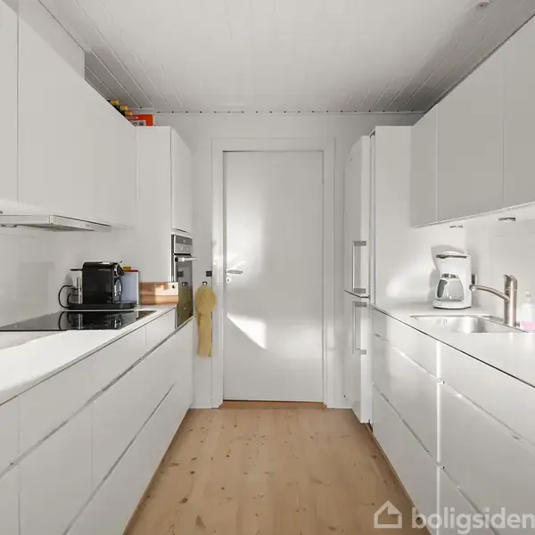 Et moderne køkken med hvide skabe og skuffer står opstillet langs begge sider af rummet. En kaffemaskine og en knivholder er synlige. Blødt naturligt lys oplyser rummet.