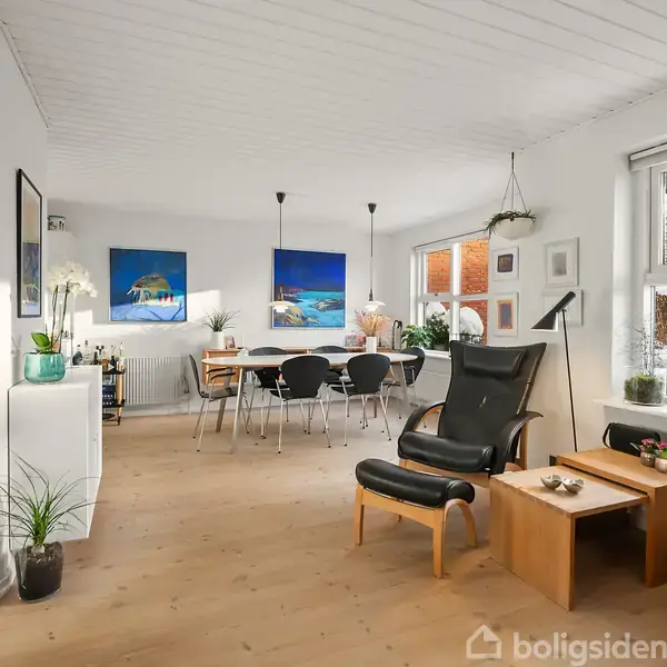 Moderne stue med sorte lænestole, sofabord og spisebord med stole. Lyse vægge, trægulv, kunstmalerier, planter og store vinduer skaber en lys og indbydende atmosfære.