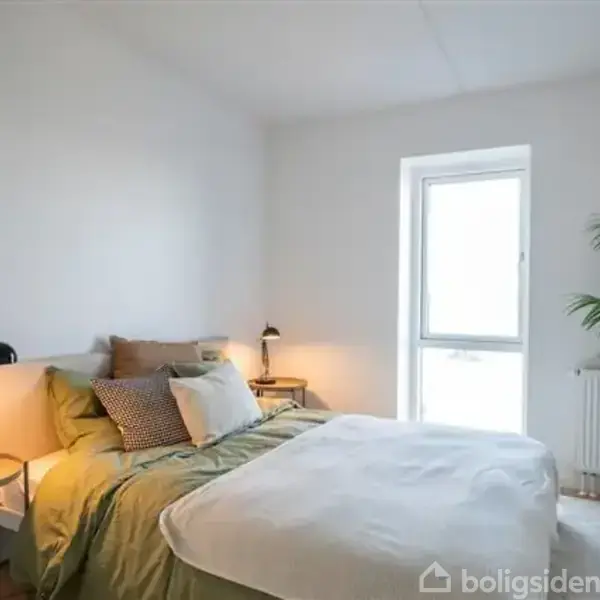 En seng med grøn sengetøj står i et lyst soveværelse. Ved siden af er to natborde med lamper. En stor plante står ved det lyse vindue.