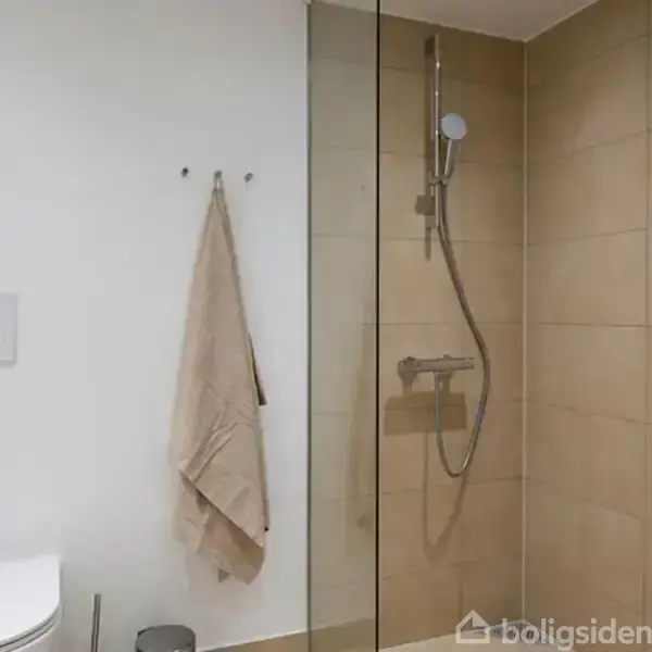 Brusekabine med håndbruser monteret på beige fliser. Ved siden af hænger et håndklæde på en krog. I forgrunden ses en væghængt toiletskål og en toiletbørste.