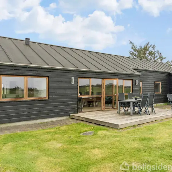 Et sortmalet hus med store vinduer vender mod en velplejet græsplæne. På terrassen står et bord med seks stole. I baggrunden ses grønne træer og en delvis overskyet himmel.