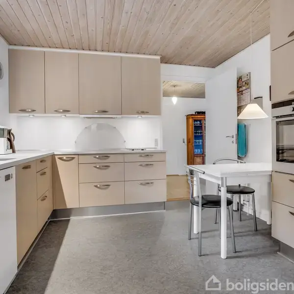 Køkken med beige skabe, opvaskemaskine og indbygget ovn, indeholder et lille hvidt bord med to sorte stole. Detaljer omfatter træloft og en kaffeautomat nær vinduet.