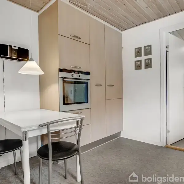 Et lille køkkenområde med et hvidt bord og to sorte stole. Et indbygget skab med ovn er i baggrunden. En gang fører til en lys større stue.