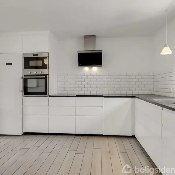 Hvidt køkken med indbyggede hvidevarer og sort bordplade. Flisevæg i baggrunden, vindue til højre. Bordlampe hænger ned fra loftet. Trægulv strækker sig gennem rummet.