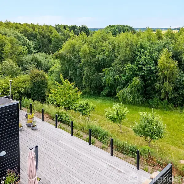 En stor terrasse af træ grænser op til en frodig skov. Et sort hus er synligt til venstre, omgivet af grønne buske og træer i en landlig beliggenhed.