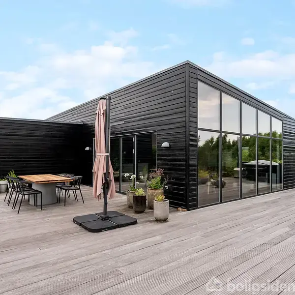 Sort, moderne hus med store vinduer står stille på en træterrasse, omgivet af planter og havemøbler. Det er en lys dag med en have i baggrunden.
