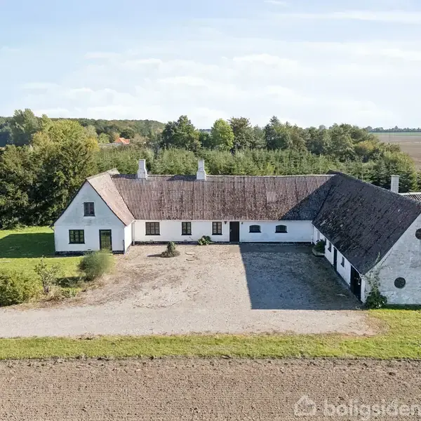 En hvid, vinkelformet landejendom står stille på en grusplads, omgivet af åbne marker og tæt bevoksning i baggrunden under en blå, skyet himmel.