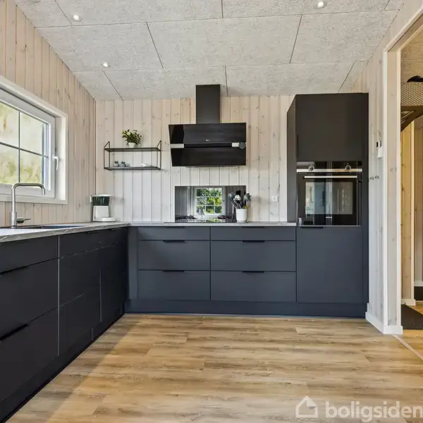Sort køkken med moderne skabe og skuffer, placeret mod en lys trævæg. Rummets trægulv og vindue giver naturligt lys. Intet tekst fundet i billedet.