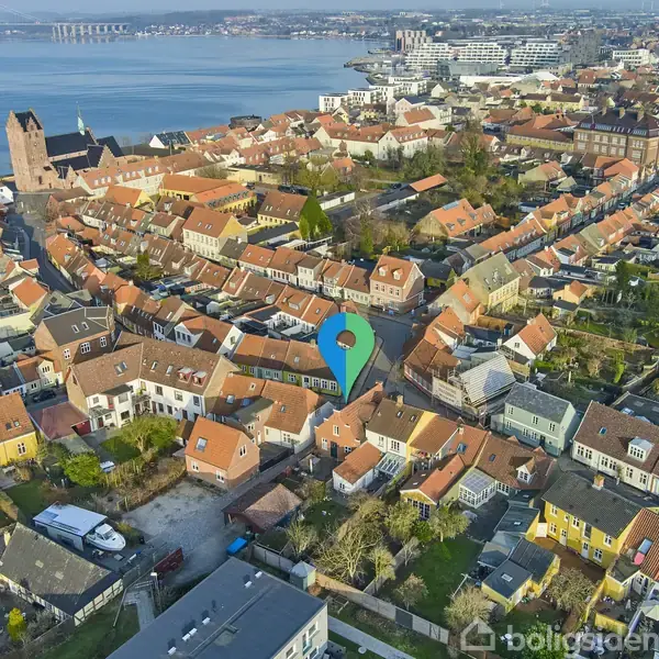 Et byområde med røde tegltage viser en tætpakket bydel set fra oven, markeret med en grøn pin. I baggrunden ses en kirke, vand og en bro.