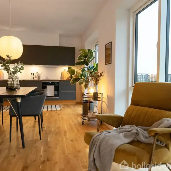 En gul lænestol med tæppe står ved siden af et stort vindue. I baggrunden er et spisebord med en lampe over, samt et køkken med mørke skabe og planter.