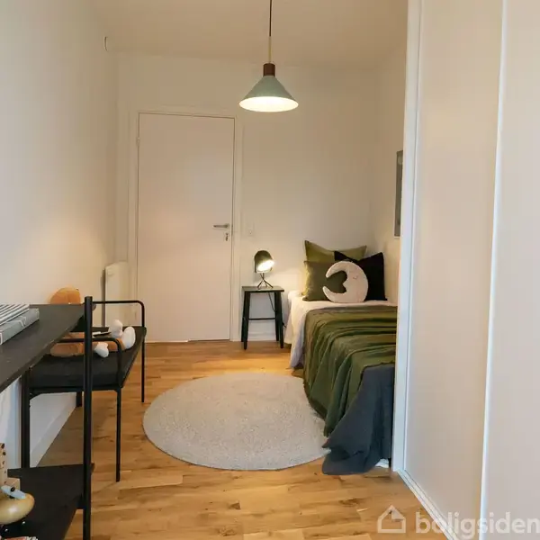 Et lille værelse har en enkeltseng dækket med en grøn plaid og puder i højre side. En rund tæppe ligger på trægulvet. Et skrivebord med lamper og bøger ses til venstre.
