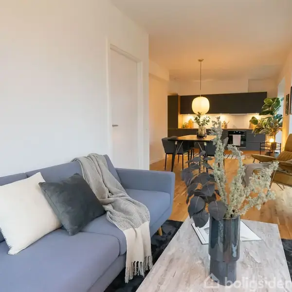 En grå sofa med puder og et tæppe står i en stue. Et sofabord med en vase er foran sofaen. I baggrunden ses en spisebordssæt og moderne indretning.