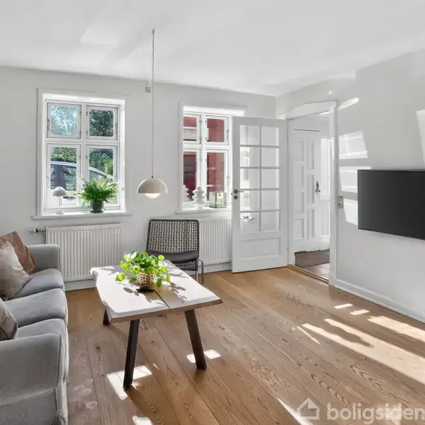 Stue med sofa og sofabord, dekoreret med planter. En fladskærm hænger på væggen, og dagslys strømmer ind gennem to vinduer. Åben dør fører til tilstødende rum.