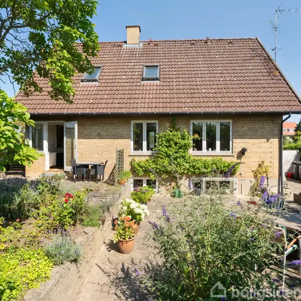 Et murstenshus med rødt tegltag, omringet af en frodig have med blomsterbede og terrasse. I forgrunden står havemøbler og en parasol. Skyfri blå himmel.