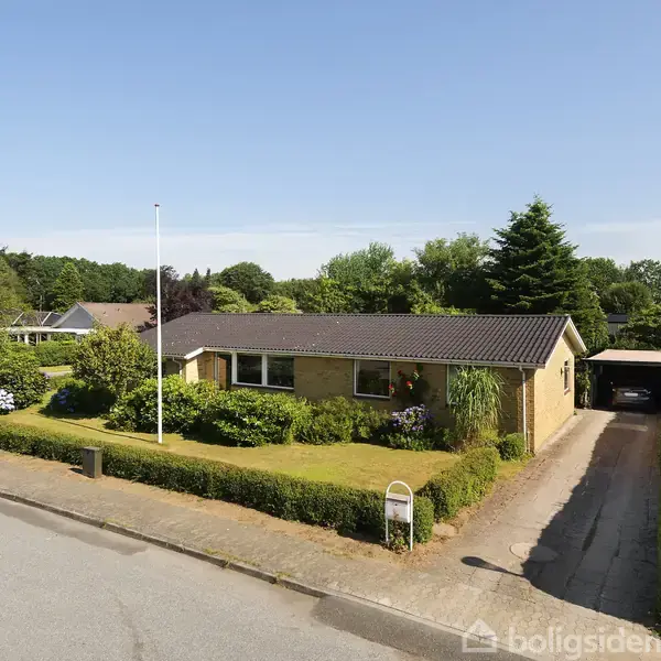 En gulstensvilla står stille omkranset af en veltrimmet have med buske og blomster. Indkørsel fører til en carport, og fleretagede træer omgiver området i et roligt boligkvarter.