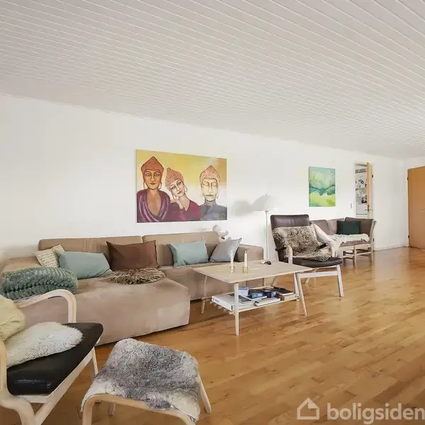 Stue med trægulv har sofaer og lænestole dækket med tæpper. Kunstværker hænger på de hvide vægge, mens spisebordet ses i baggrunden. Klar belysning og minimalistisk indretning.
