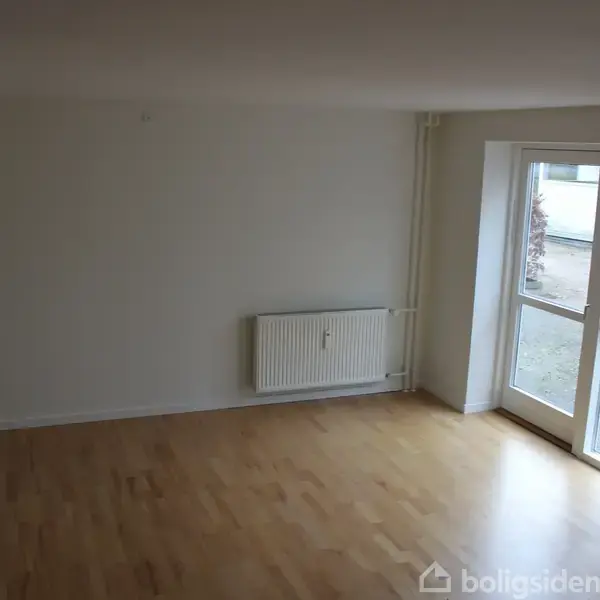 Et tomt værelse med trægulv har et vindue og en dør ud mod en grusbelagt gård. Der er en radiator monteret på væggen. Ingen tekst til stede.