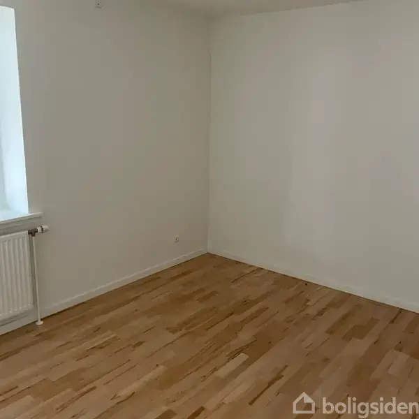 Et tomt rum har lyse trægulve og hvide vægge. En radiator er placeret ved et vindue til venstre, hvor noget lys kommer ind.