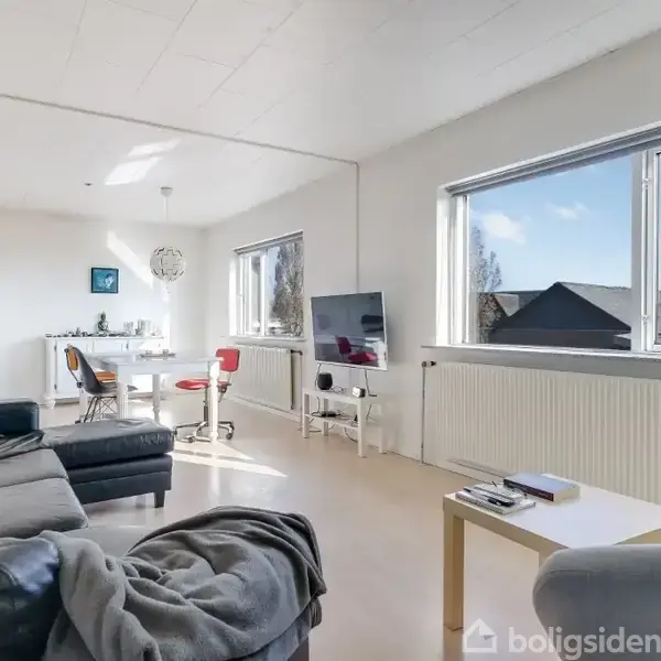 Sofa står foran et sofabord i en lys stue med store vinduer. I baggrunden er der et spisebord med stole. Tv'et står ved siden af vinduet.