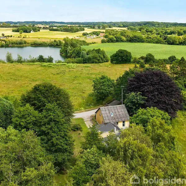 Et hus står omgivet af tætte træer. En vej løber ved siden af huset. I baggrunden ses en sø og marker under en blå himmel i et landligt område.