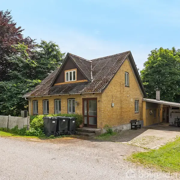 Gult murstenshus står på en grusvej, omgivet af grønne træer. Huset har skråtag og en carport. Tre skraldespande står ved siden af. Skilt på hegnet: "VILLA TIL SALG 139 M2".