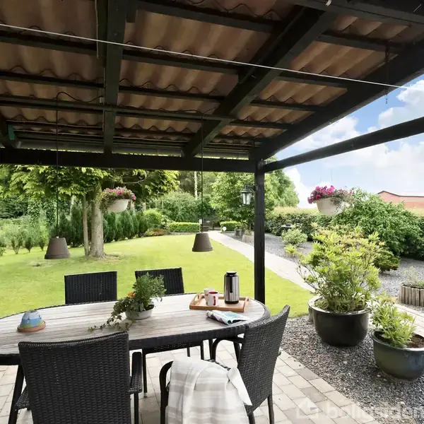 Et ovalt havebord med stole står under en pergola. Bordet har en termokande og små dekorationer. Omgivet af en velplejet græsplæne og buske i en baghave.