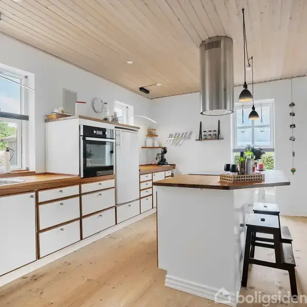Et lyst køkken med hvidt inventar og træoverflader. Et moderne komfur og vask er på en række, med store vinduer, der giver naturligt lys. Bardisk med høje stole.
