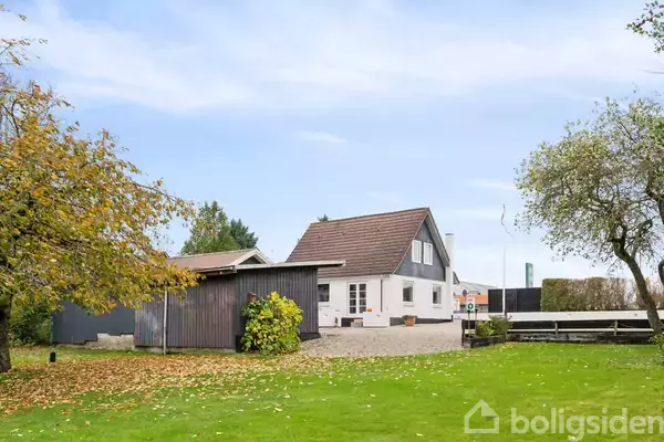 Et hvidt hus med rødt tag står i en grøn have med et træ til venstre. Et sort udhus er ved siden af huset. Himlen er blå med lette skyer.