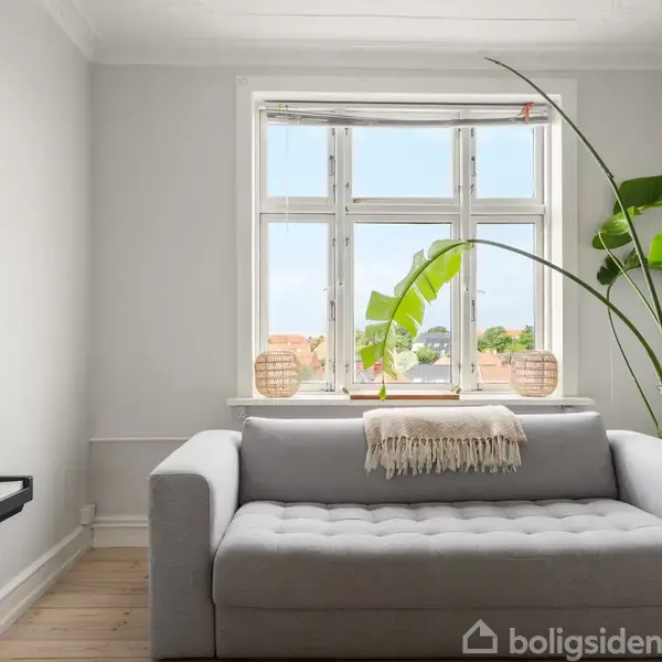 Sofa står i en lys stue foran et vindue med udsigt over hustage. En stor potteplante står ved siden af, og et el-klaver er placeret til venstre for sofaen.