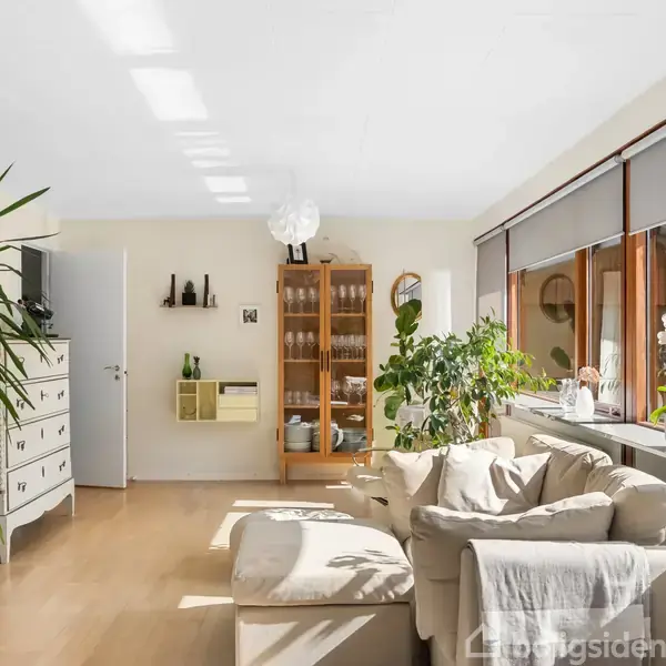 Stue med beige sofa og grønne planter ved et stort vindue. En hvid kommode og et vitrineskab står langs væggen. Sollys strømmer ind gennem vinduerne og oplyser rummet.