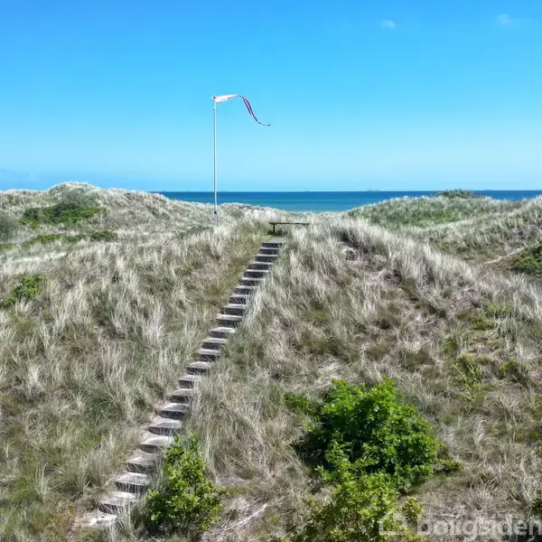 Trappetrin fører op ad en græsklædt klit med en flagstang på toppen, omgivet af blå himmel og hav i baggrunden.