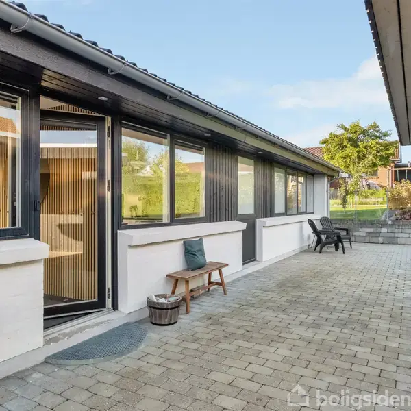 En hvid murstensbungalow med sort tag står i sollys. Foran huset er en stenbelagt sti omgivet af grønne buske og græsplæne under en klar blå himmel.
