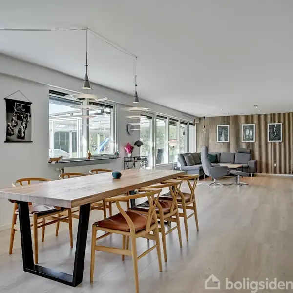 Et langt, hvidt hus med sort tag har store vinduer. Til højre er der en brolagt gangsti og et sort skur. Blå himmel og planter i baggrunden.