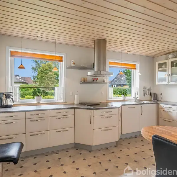 Et lyst køkken har hvide skabe og grå bordplader, en køkkenø med kogeplade, en emhætte, og to vinduer med udsigt til træer. Der er en sort stol og en rund træbordplade.