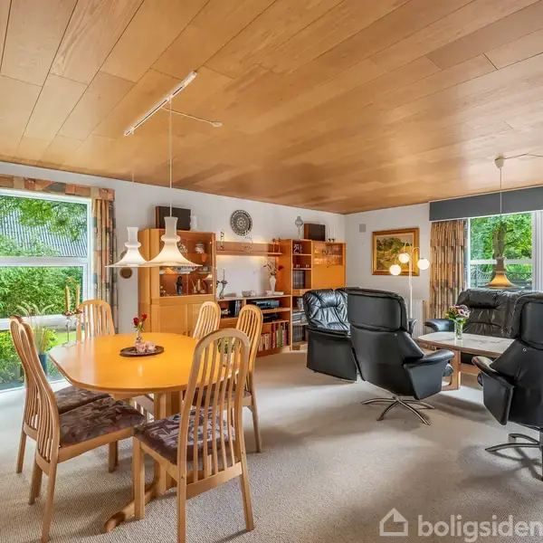 Et lyst opholdsrum med træloft, indeholder et spisebord med stole i centrum og to sorte lænestole til højre. Store vinduer giver udsigt til grønne buske udenfor.