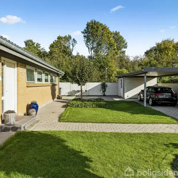 En bil parkerer i en carport ved siden af en gul murstensbygning. Foran bygningen er en velplejet græsplæne og flisebelagt gangsti. Træer omgiver gårdspladsen.