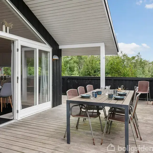 Terrassebord opdækket til seks personer med bestik og glas. Bordet står på en træterrasse ved et moderne, sort-hvidt hus med åbne døre til stue. Grønne træer omgiver området.