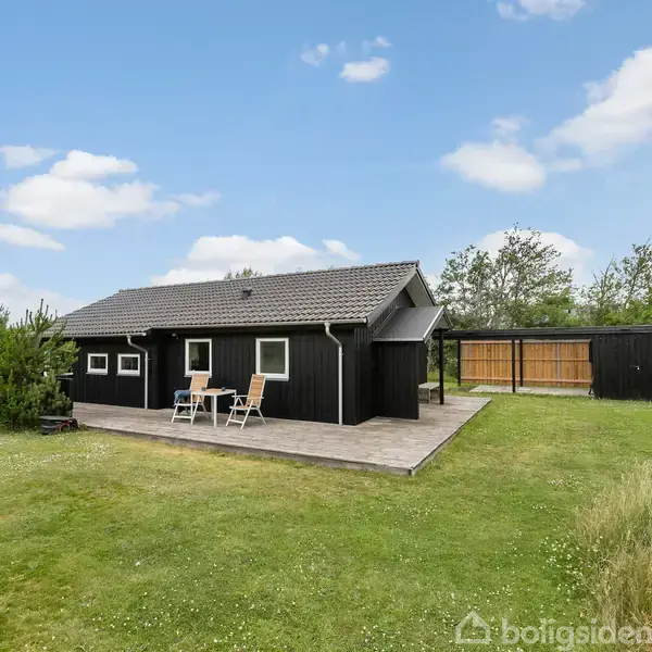 Sommerhus med sort træfacade står stille på en græsplæne, omgivet af buske og træer. To stole og et bord er placeret på en terrasse foran huset under en blå himmel med skyer.