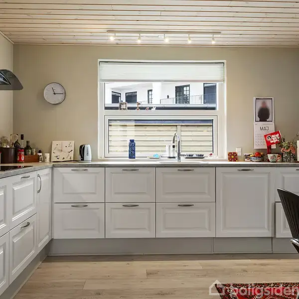 Hvidt køkken med hvide skabe fyldt med genstande. Vinduet har udsigt til nabohuse. Ur på væggen viser klokken. Bord med krus og køkkenredskaber. Plakat med teksten "MÅL".
