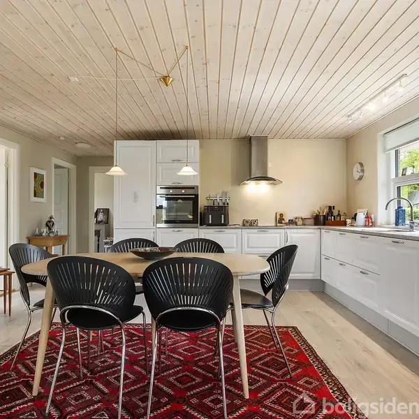 Et moderne køkken-alrum med et spisebord og seks sorte stole. Bordet står på et rødt mønsteret tæppe. Rummet er lyst med træloft og store vinduer.