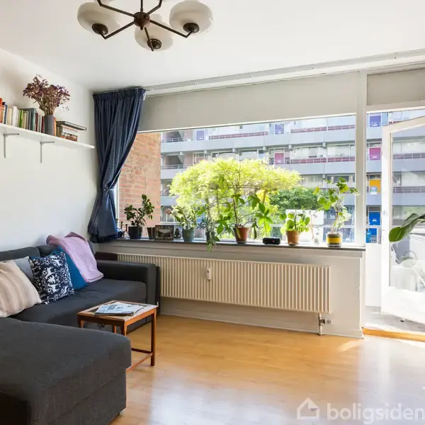 En stue med en grå hjørnesofa fyldt med farverige puder. På en hylde over sofaen står bøger og planter. Store vinduer med udsigt til altaner.