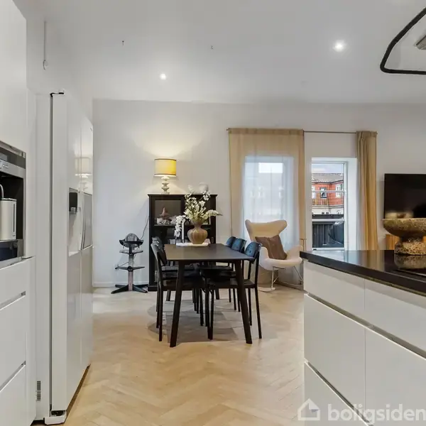 Køkken-alrum med hvide skabe og sort bordplade. Spisebord med stole centralt placeret, omgivet af en lampe og pyntegenstande. Store vinduer giver udsigt til en udendørs terrasse.