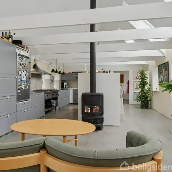 En moderne stue med rund sofa og træbord foran en brændeovn. Køkkenet i baggrunden har grå skabe og en stor komfur. Vægge prydes med grønne planter og en rund vægur.
