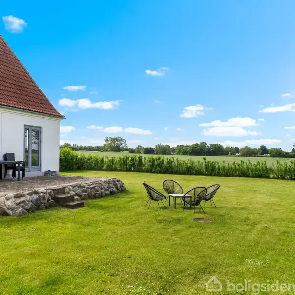 Et hvidt hus med rødt tag har en terrasse med sorte møbler. Foran er en græsplæne med et cafébord og stole. Landskabet er grønt og åbent under en blå himmel.