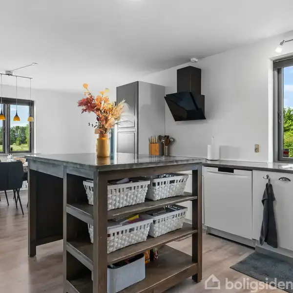 Køkken med centralt placeret køkkenø fyldt med kurve, omkranset af moderne hvide skabe. Spisebord med stole i baggrunden nær vinduer, hvor sollys skinner ind og udsigt til grønne omgivelser ses.