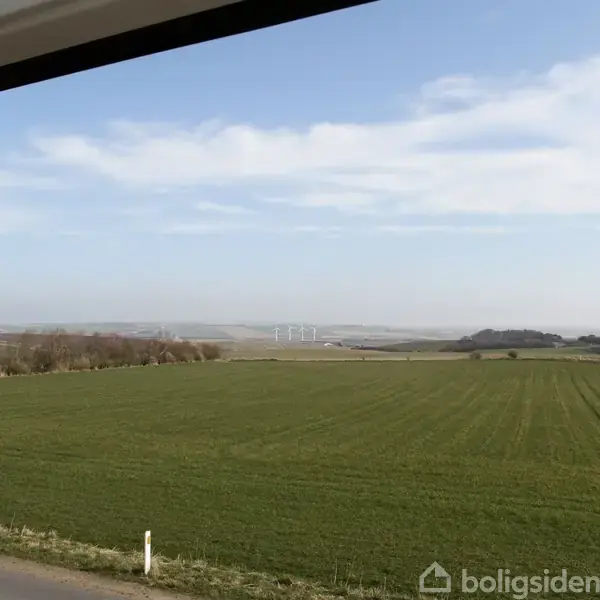 Grøn mark strækker sig mod horisonten under en klar blå himmel; i det fjerne ses vindmøller og en sø, omkranset af spredte træer og buske.
