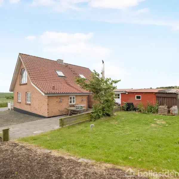 Et murstenshus med rødt tag står stille på en græsplæne ved en grusvej i landlige omgivelser med marker i baggrunden.