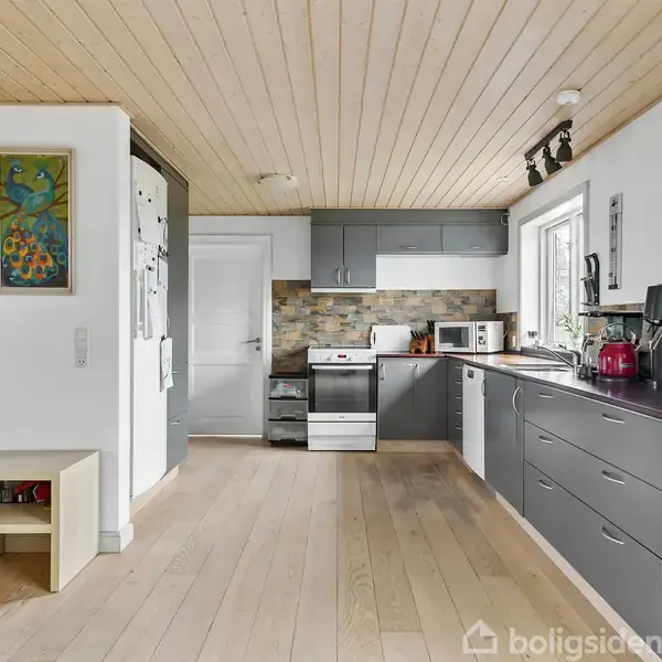 Et moderne køkken med grå skabe og hvide vægge. Der står en ovn og mikroovn på køkkenbordet, samt diverse små apparater. Et vindue lader naturligt lys ind i rummet.